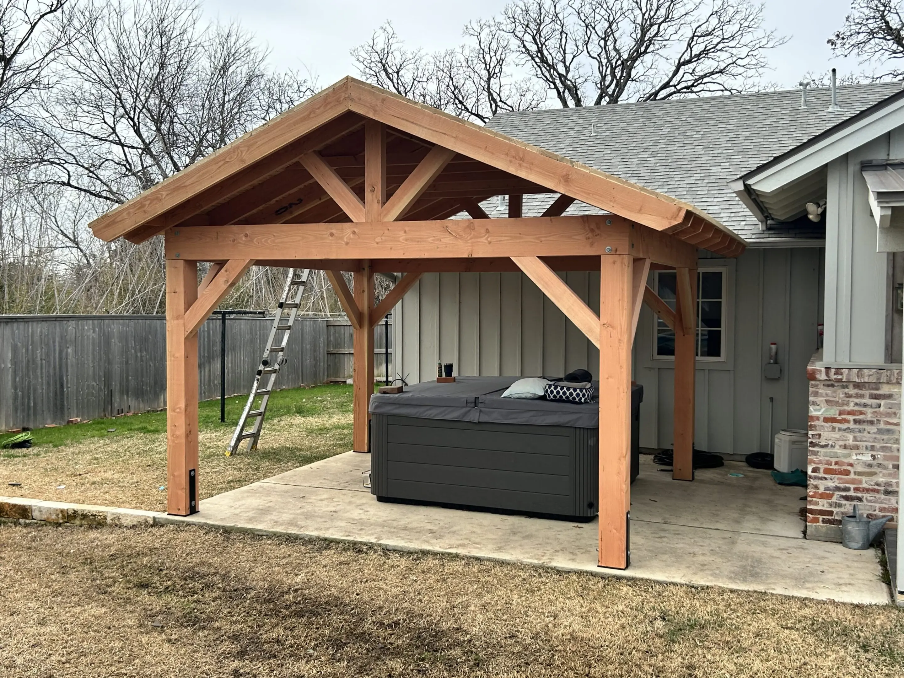 Wooden pergola sheltering a hot tub on a concrete patio beside a house.
