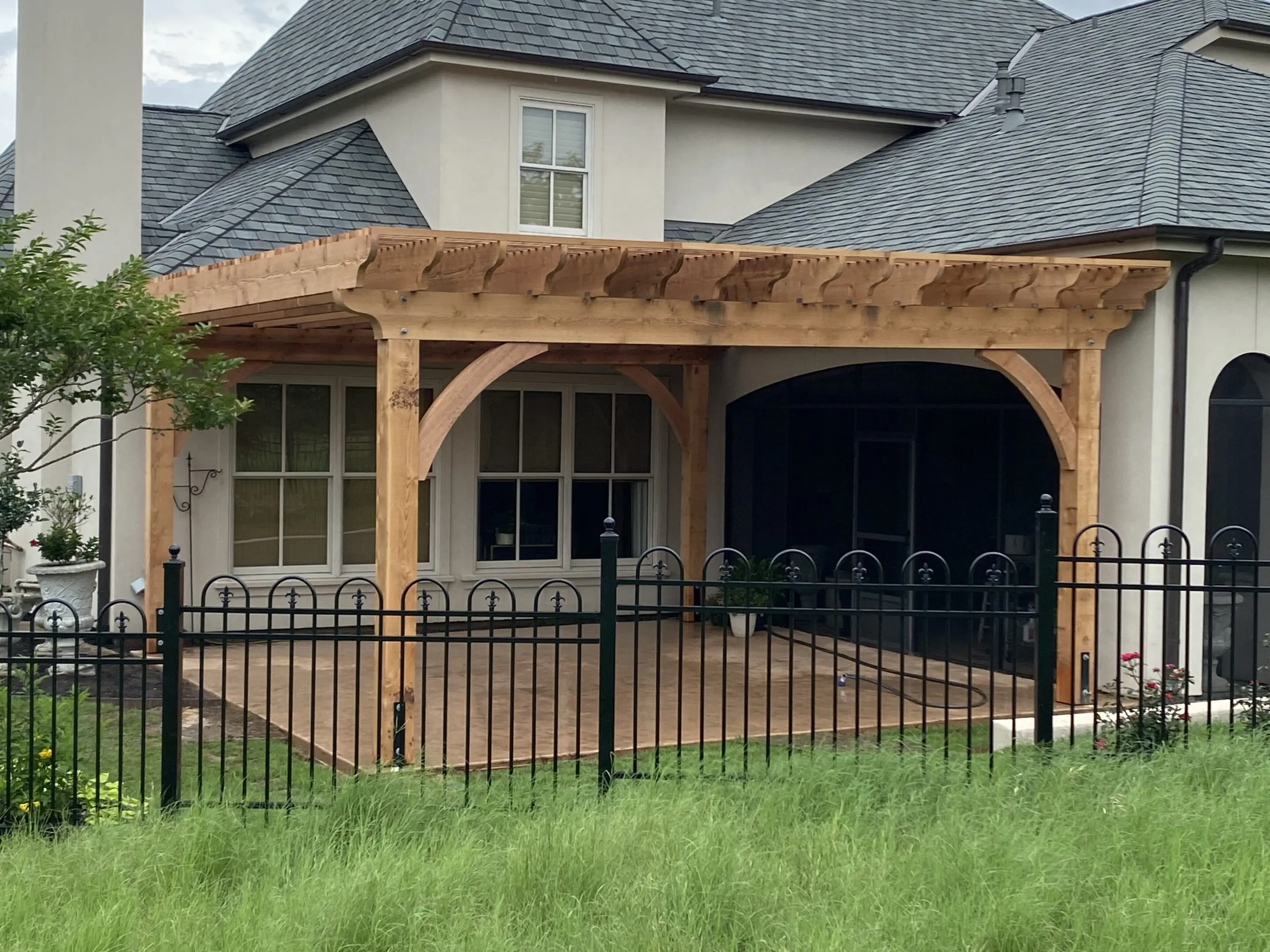 Wooden pergola attached to a house with a metal fence in front.